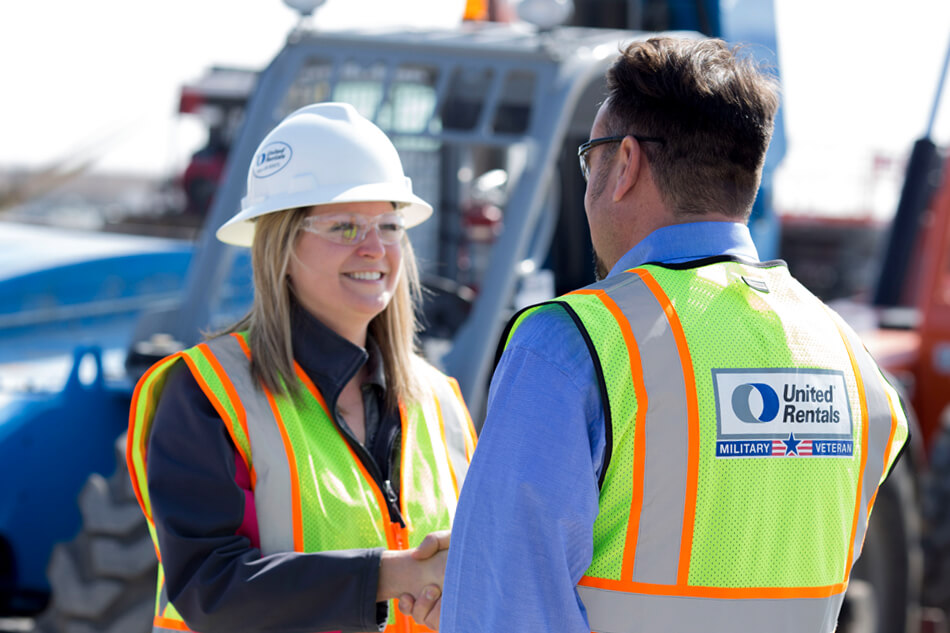 A smiling female worker in a hard hat and a yellow vest talks to a male colleague at a construction site.
